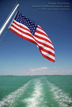 Picture of an American flag hanging off the back of a large boat; wake behind boat is visible in the inviting blue-green water.  Find more great free photos and wallpapers at http://www.FreePhotoCourse.com. © 2011, FreePhotoCourse.com; all rights reserved. 
